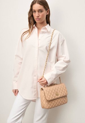 Sac à main beige matelassé avec une bandoulière chaîne dorée, tenu contre une chemise rose clair boutonnée et un pantalon blanc. Texture lisse, fermeture à rabat.