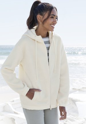 Cream plush fleece zip-up hoodie with a drawstring hood, front pockets, and ribbed cuffs, layered over a grey and white striped shirt.