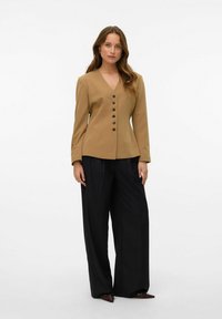 Blazer ajusté marron clair avec un col en V, six boutons noirs, et des manches longues, associé à un pantalon noir ample et des chaussures pointues.