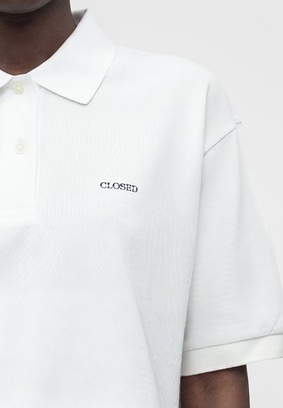 Λευκό πόλο με κοντά μανίκια, γιακά και δύο κουμπιά, με μικρό μαύρο κεντημένο κείμενο "CLOSED" στο αριστερό στήθος.