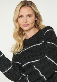O femeie blondă cu păr ondulat poartă un pulover tricotat negru cu dungi albe, privindu-ne direct cu o expresie neutră pe un fond deschis.
