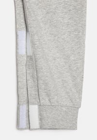 Pantalons de sport grisés avec une texture lisse, des poignets élastiques et deux accents en Velcro blancs sur le côté pour un ajustement réglable.