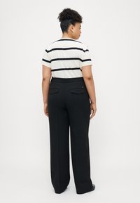 Mustat leveälahkeiset housut, joissa on sileä pinta ja kaksi takataskua. Yhdistetty valkoisiin ja mustiin raidallisiin lyhythihaisiin paitoihin.