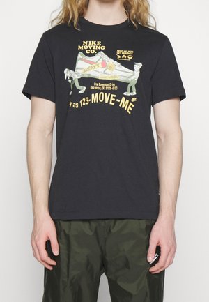 T-shirt noir avec illustration de deux personnages de dessin animé portant une grande chaussure de sport étiquetée "lourde" et texte promouvant Nike Moving Co.