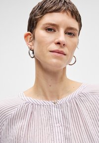 Ανοιχτό ροζ πουκάμισο με ρίγες, στρογγυλή λαιμόκοψη, κούμπωμα με κουμπιά και συγκεντρωμένες λεπτομέρειες. Το μοντέλο φοράει ασημένιες κρίκους σκουλαρίκια.