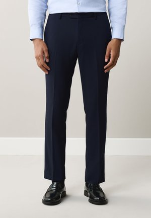Homme portant un pantalon de costume bleu marine foncé, une chemise bleu clair et des chaussures en cuir noir, debout devant un fond uni.