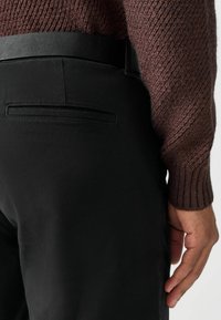 Fekete nadrág sima textúrával, átlapolt zsebbe és fekete övvel. A tetején részben látható egy barna kötött pulóver.