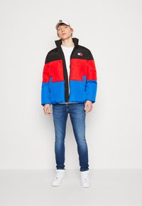 Giacca a vento con blocchi di colore neri, rossi e blu, chiusura frontale con cerniera e dettaglio del logo. Indossata sopra una maglietta bianca e abbinata a jeans blu.