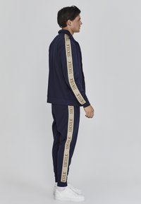 Námornícky športový komplet s béžovými bočnými pruhmi, na ktorých je názov značky "SIKSILK". Mäkko tkaný materiál, zavinovací kabát a zodpovedajúce nohavice s rebrovými manžetami.
