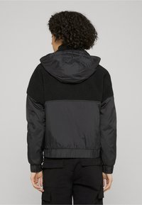 Chaqueta negra con textura contrastante en la parte superior de la espalda con material tipo forro polar. Incluye capucha y puños y dobladillo elásticos.