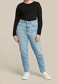 Femme portant un haut à manches longues noir, un jean taille haute bleu clair et des baskets blanches, debout sur un fond beige uni.