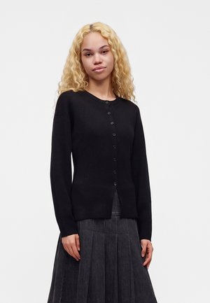 Jeune femme aux longs cheveux blonds bouclés porte un cardigan noir boutonné et une jupe plissée sombre sur un fond clair uni.