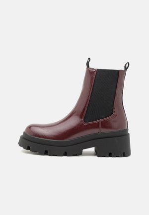 Botas de tobillo de charol burdeos con suela negra gruesa, paneles laterales elásticos y lengüetas de tirón, con un diseño robusto y voluminoso.