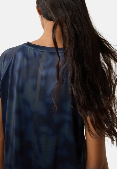 Frau mit langem dunklem Haar, die ein dunkelblaues, gemustertes Sportshirt trägt, von hinten gezeigt.