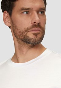 Balts crew-neck T-krekls ar gludu tekstūru. Parāda tuvplānu skatu uz vīrieša kaklu un žokļa līniju, redzama stublas un neitrāla izteiksme.
