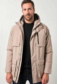 Uomo che indossa un parka beige sopra un maglione nero a trecce e jeans scuri, in piedi contro uno sfondo chiaro e semplice.