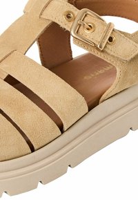 Béžová semišová sandála se širokými pásky a přezkou. Má opálenou stélku a tlustou, texturovanou béžovou podešev pro stabilitu.