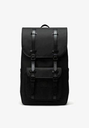 Sac à dos en tissu noir avec un rabat supérieur, deux sangles à boucle et une poche avant. Présente une surface texturée et un logo de marque cousu.