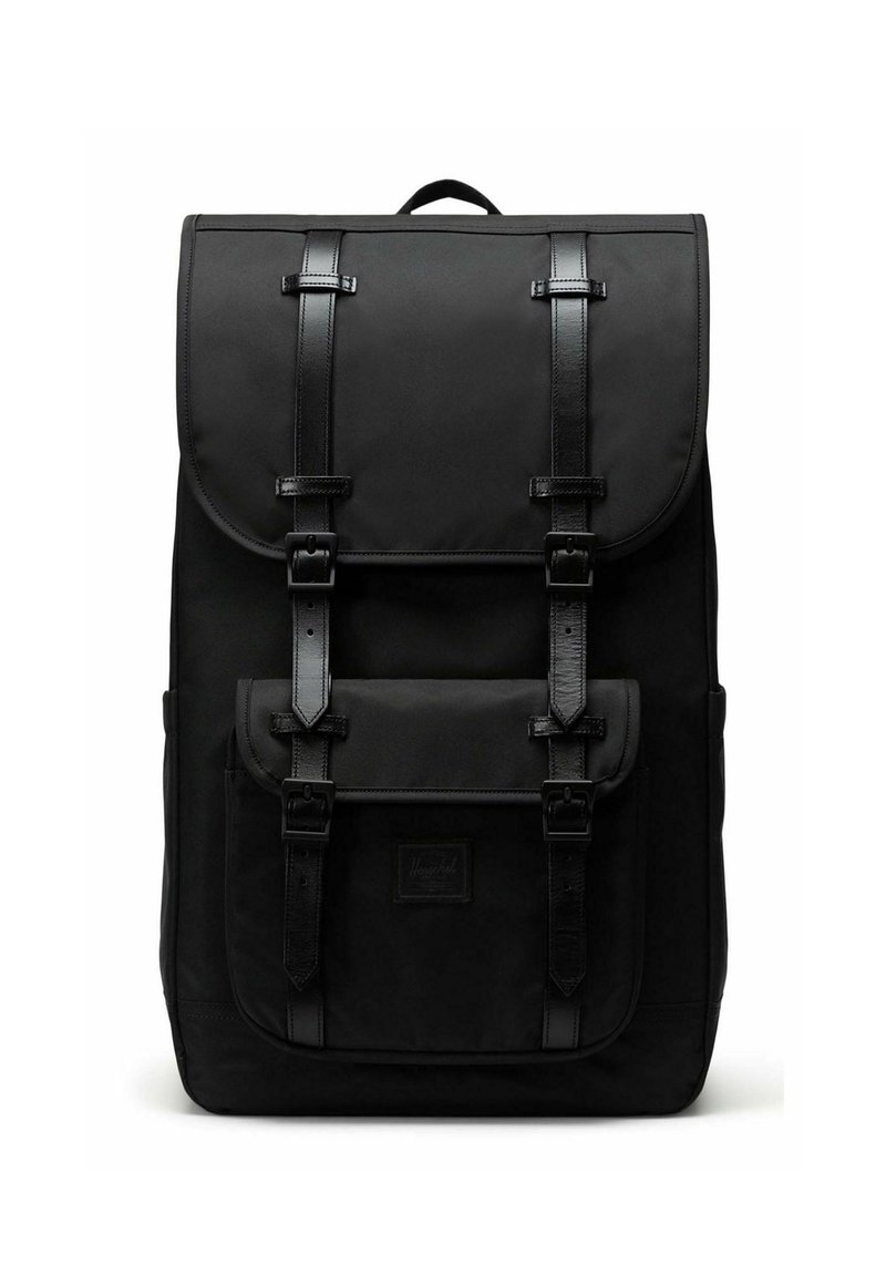 Sac à dos en tissu noir avec un rabat supérieur, deux sangles à boucle et une poche avant. Présente une surface texturée et un logo de marque cousu.