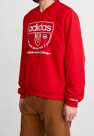 Sweatshirt rouge à capuche arborant un grand logo Adidas blanc et du texte. Manches longues avec des poignets élastiques. Fabriqué en tissu lisse.