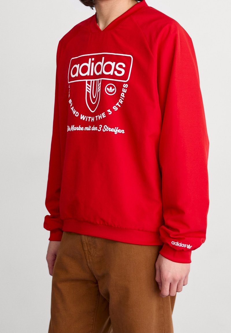 Red pullover sweatshirt featuring a large white Adidas logo and text. Long sleeves with elastic cuffs. Made of smooth fabric.