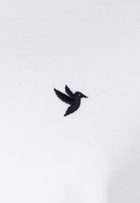 Tela blanca con un pequeño logotipo de colibrí negro bordado en el lado izquierdo. Textura suave, diseño clásico.
