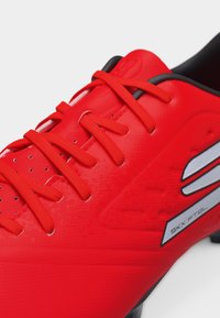 Εγγύς πλάνο κόκκινου ποδοσφαιρικού παπουτσιού με κόκκινες κορδόνια και ασημένιο λογότυπο στο πλευρό, με μαύρο γιακά και καρφιά στη σόλα.