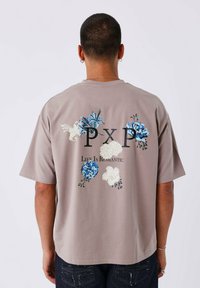 Camiseta gris de manga corta con embellishments florales en azul y blanco, con el texto "PXP La Vida Es Romántica" en negro.
