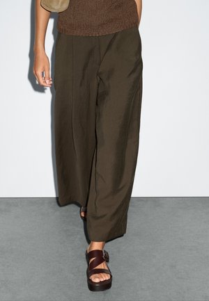 Brede, donkergroene broek van een gestructureerde stof, gecombineerd met bruin leren sandalen met een gespsluiting. Bruine gebreide top zichtbaar.