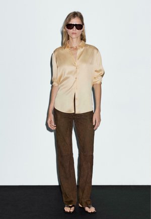 Satin, beige shirt met korte mouwen en knoopdetails, gecombineerd met bruine suède rechte broek. Het model draagt donkere zonnebril en minimalistische sandalen.