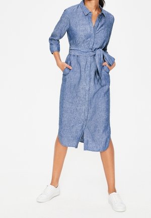 Frau trägt ein blaues Hemdblusenkleid mit langen Ärmeln, Taillengürtel und Fronttaschen, kombiniert mit weißen Turnschuhen, steht mit den Händen in den Taschen.