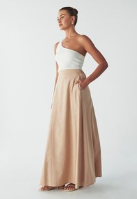Gonna maxi beige chiaro in tessuto fluido con tasche laterali, abbinata a un top bianco a coste con spalla scoperta. Texture liscia, drappeggio elegante.