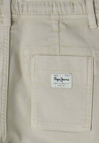Beige bomullbyxor med en bakficka som har en vit etikett med texten "Pepe Jeans", sydda kanter och en mjuk textur.