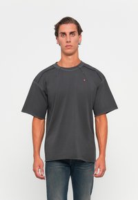 Young man with light skin and short brown hair wearing an oversized faded black T-shirt and blue jeans, standing against a white background.