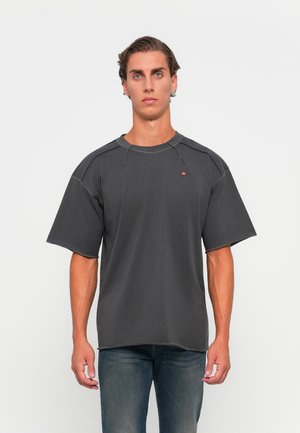 Young man with light skin and short brown hair wearing an oversized faded black T-shirt and blue jeans, standing against a white background.