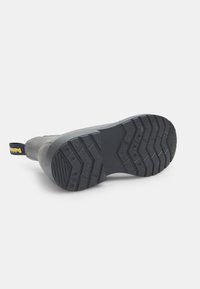 Bota con suela de goma, parte superior texturizada de color gris y una lengüeta amarilla y azul marino. Presenta un patrón de banda de rodadura en zigzag para mayor tracción.