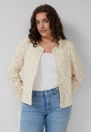 Kobieta z kręconymi włosami, ubrana w kremową koronkową kurtkę na białym topie i niebieskich dżinsach, stoi na jednolitym jasnym tle.