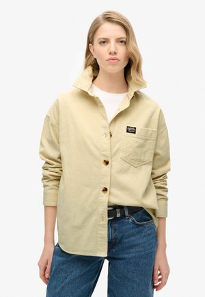 Chemise en velours côtelé kaki avec col, boutons sur le devant, poche plaquée et étiquette de marque. Portée avec un jean bleu, présentant une finition texturée.