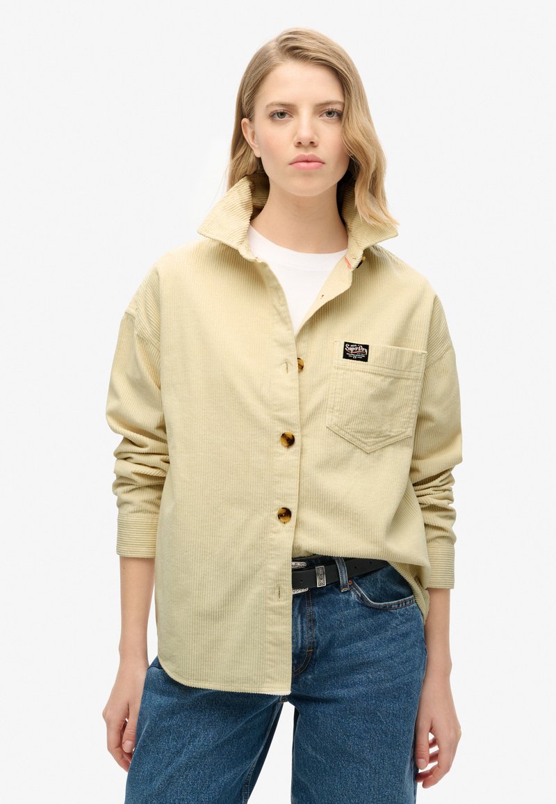 Chemise en velours côtelé kaki avec col, boutons sur le devant, poche plaquée et étiquette de marque. Portée avec un jean bleu, présentant une finition texturée.