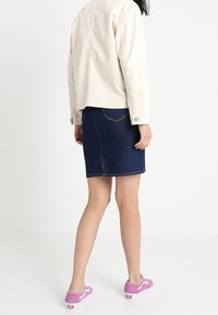 Person som står klädd i en krämfärgad jacka, en mörkblå denimkjol med slits bak, och rosa sportiga sneakers mot en vit bakgrund.