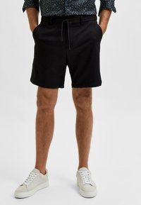 Černé šortky s uvolněným střihem, s páskem na šňůrku a bočními kapsami, spárované se světle šedými teniskami s bílými tkaničkami a hladkým povrchem.