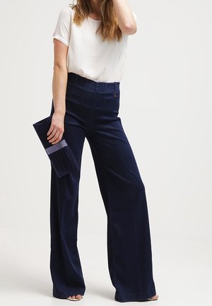 Femme portant un haut blanc à manches courtes et un pantalon large taille haute bleu marine, tenant une pochette bleu marine ornée d'une bande de strass.