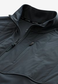 Chaqueta activa de media cremallera de color gris oscuro con textura suave, paneles acolchados y bolsillo en el pecho con cremallera. Presenta un cuello alzado y puños elásticos.