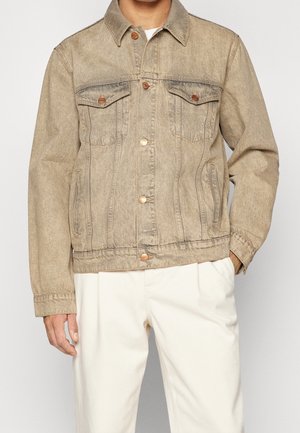 Mężczyzna ubrany w beżową kurtkę jeansową z zapinanymi na guziki kieszeniami na piersi oraz białe spodnie, stoi z jedną ręką w kieszeni.