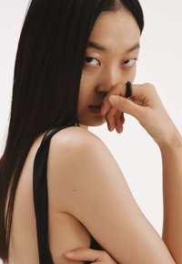 Top sin mangas negro con un escote profundo. Primer plano de una persona con el cabello liso y oscuro, sosteniendo su mano sobre la boca.