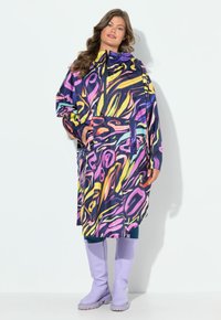 Femme portant un imperméable à motif abstrait multicolore avec capuche, se tenant les mains dans les poches, vêtue de bottes hauteur genou de couleur lilas.