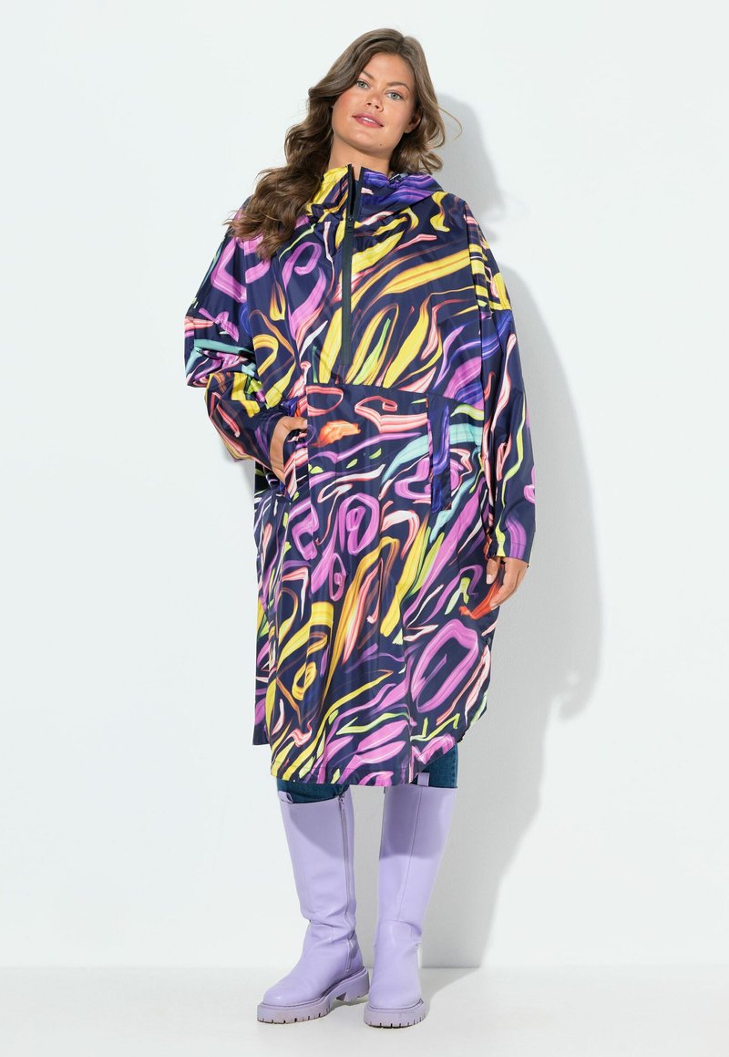 Donna in un impermeabile con motivo astratto multicolore e cappuccio, in piedi con le mani nelle tasche, indossando stivali al ginocchio di un leggero viola.