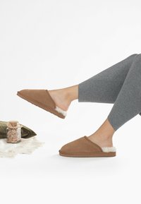 Bruna mockaskor med en fluffig vit foder, öppen baksida, platt gummisula och minimalistiska sömnadsdetaljer.