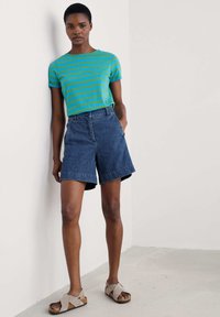 Camiseta de manga corta a rayas en tonos azul y verde teja, combinada con pantalones cortos de mezclilla de tiro alto. La modelo lleva sandalias grises de toe abierto. Fondo neutro.