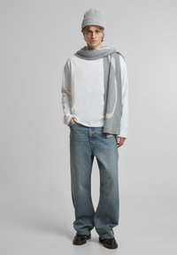 Weißes Langarmhemd, blaue locker sitzende Jeans, graue Strickmütze, grauer Schal mit einem hellen Muster, schwarze Schuhe. Schlichter Hintergrund.